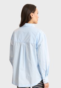 Camicia oversized azzurra chiaro con piega sulla schiena, maniche lunghe, orlo arrotondato e bottoni ai polsini. Tessuto morbido e leggero.
