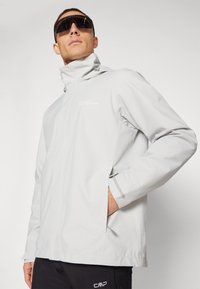 Chaqueta impermeable de color gris claro con cuello alto, cierre de cremallera y un detalle sutil del logo. Cuenta con puños ajustables y bolsillos laterales.
