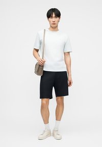 Jeune homme debout portant un t-shirt gris clair, un short noir, des chaussettes blanches, des baskets beiges et un sac bandoulière taupe.