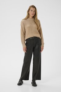 Woman standing with long blonde hair wearing a beige sweater, black wide-leg leather pants, and black loafers against a plain background.