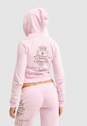 Persoon draagt een roze Juicy Couture velours trainingspak met capuchon omhoog, met logo en tekst op de rug en linkerbeen.