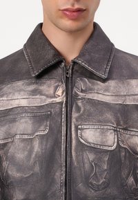 Izbledela siva denim jakna z zadrgo, s koničastim ovratnikom, dvema žepoma na prsih in subtilnimi teksturnimi odstopanji na tkanini.