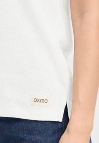 Top bianco con texture e una leggera lucentezza, con etichetta visibile che recita "OXMO", abbinato a jeans in denim blu scuro.