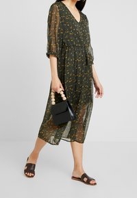 Vestido floral verde con escote en V, material transparente y mangas tres cuartos. Combinado con un bolso negro y sandalias marrones tipo slide.