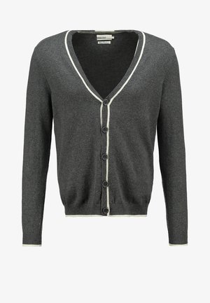 Mann trägt graue, geknöpfte Strickjacke über weißem T-Shirt, schwarze Hose und weiße Slip-On-Schuhe, steht vor schlichter Hintergrund.