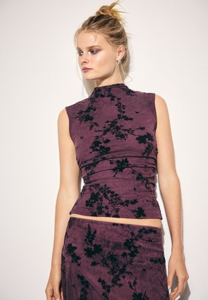 Giovane donna con capelli biondi raccolti in uno chignon, indossa una canotta viola senza maniche e una gonna abbinata con motivi floreali neri, in piedi contro un muro bianco.