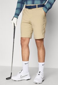 Pantalones cortos de golf beige con un diseño clásico, combinados con un cinturón azul marino. Zapatillas de golf blancas con detalles negros y calcetines blancos hasta el tobillo. Palo de golf en la mano.