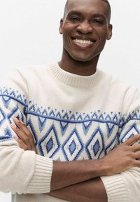 Vaalea kermanvärinen neulepusero, jossa on sininen geometrinen timanttikuosi, neulottu kangas ja resorit hihansuissa. Malli hymyilee ja pitää käsiään ristissä.