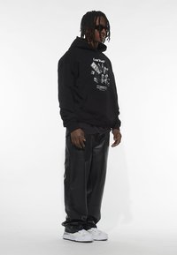 Crna kapuljača s grafičkim printom, u kombinaciji s labavim crnim kožnim hlačama i bijelim teniscima s teksturiranim detaljima.