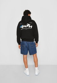 Sort hættetrøje med "YOUTH" grafisk tryk i hvid og blå på ryggen, parret med blå denimshorts og hvide sneakers med blå detaljer.