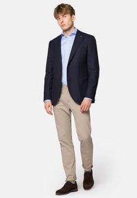 Granatowa marynarka z dwoma guzikami, jasnoniebieska koszula, beżowe spodnie i brązowe zamszowe buty. Casułowy, ale elegancki wygląd z dopasowanym krojem.