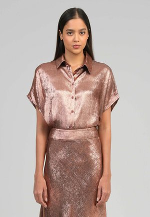 Femme aux longs cheveux foncés portant une chemise boutonnée rose doré brillante et une jupe taille haute assortie, devant un fond gris uni.