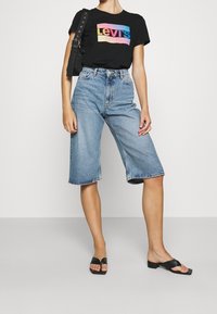 Mustapainoinen graafinen t-paita, jossa on monivärinen "Levi's"-logo, yhdistettynä vaaleansinihin denim-culotteihin, joissa on raakareuna, ja mustiin liukutossuihin.