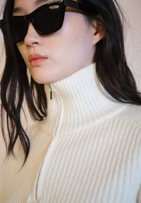 White ribbed sweater with high collar, featuring a silver zipper pull. Black sunglasses with angular design frame the face. Smooth, dark hair.