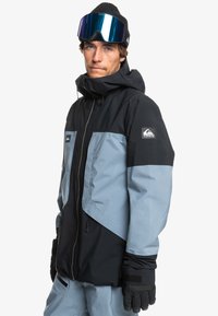Černá a světle modrá lyžařská bunda s kapucí, zapínáním na zip a nastavitelnými manžetami. Materiál se zdá být nepromokavý; logo nášivka na rukávu.