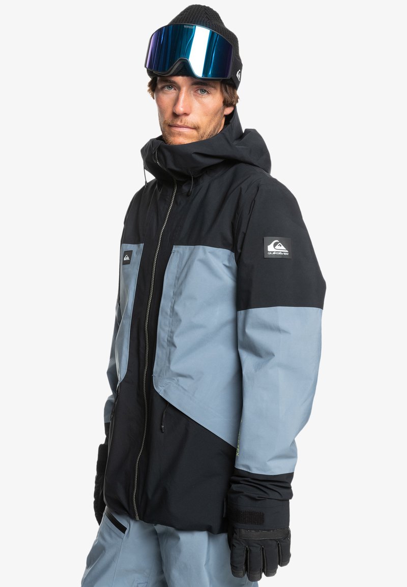 Czarna i jasnoniebieska kurtka narciarska z kapturem, zamkiem błyskawicznym i regulowanymi mankietami. Materiał wydaje się wodoodporny; naszywka z logo na rękawie.
