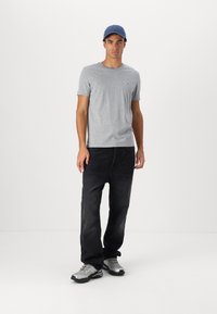 Tricou gri cu mânecă scurtă, blugi închis și adidași gri. Căciula albastră are un mic logo. Un outfit simplu și casual pe un fundal alb.
