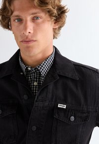 Ung mand med krøllet hår iført en sort Wrangler-jakke over en sort-hvid ternet skjorte, ensartet lyst baggrund.