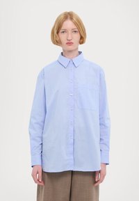 Camicetta azzurra chiara con colletto, maniche lunghe e taschino sul petto. Realizzata in tessuto liscio con una vestibilità leggermente ampia.