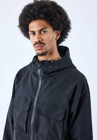 Giubbotto leggero nero con chiusura a zip, cappuccio e quattro tasche frontali; presenta un tessuto testurizzato e dettagli del logo discreti.