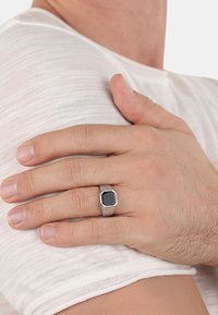 Anello esagonale in tonalità argento con un centro in pietra nera testurizzata. La mano poggia delicatamente su una maglietta bianca e testurizzata. Toni della pelle sottili visibili.