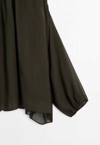 Tissu transparent brun foncé avec une manche longue ample et flottante resserrée au niveau du poignet, suspendue sur un fond blanc uni.