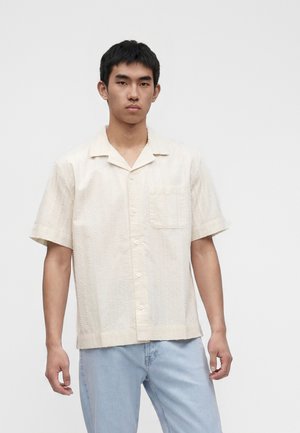 Jeune homme portant une chemise crème texturée à manches courtes et un jean bleu clair, debout devant un fond blanc uni.