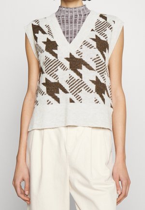 Gilet sans manches à col en V avec motif géométrique marron et beige, superposé à un col roulé côtelé taupe, associé à un pantalon crème taille haute.