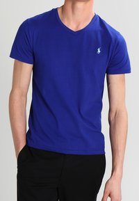 Man som bär en enkel kunglig blå V-hals T-shirt med en liten vit logotyp på vänster bröst, tillsammans med svarta byxor.