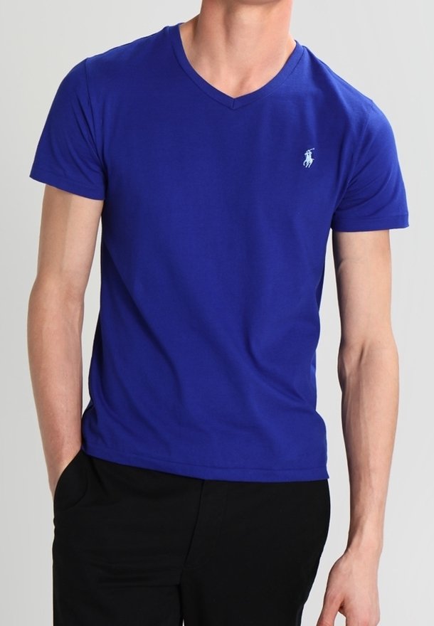 Man som bär en enkel kunglig blå V-hals T-shirt med en liten vit logotyp på vänster bröst, tillsammans med svarta byxor.