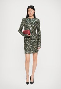 Sjajna zelena i zlatna haljina s weekama i dugim rukavima, ukrašena shoulder pads, u kombinaciji s crvenom clutch torbicom i crnim cipelama na špic.