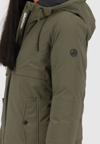 Parka jacket verde oliva con cappuccio nero, coulisse regolabile, zip frontale e tasche laterali. Presenta un accento con il marchio sul petto.
