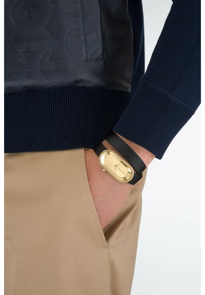 Marineblauer Pullover mit einer strukturierten Lederpasse, braune Hose und eine goldfarbene Uhr mit schwarzem Armband, die eine stromlinienförmige ovale Form zeigt.