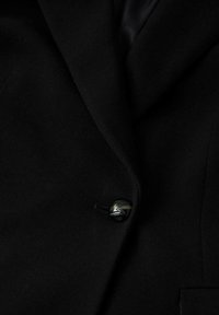 Blazer negro con una tela suave, que presenta un botón destacado y solapas con muesca. El diseño general es entallado y estructurado.