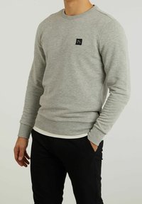 Homme portant un sweat-shirt gris clair à col rond avec un petit patch logo noir et un pantalon noir, une main dans la poche.