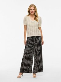 Top beige a maniche corte in maglia con scollatura a V; pantaloni neri a gamba larga con un motivo geometrico bianco; modella che indossa sandali chiari.