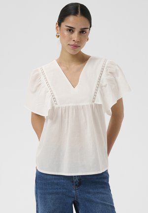 Frau mit dunklen Haaren, die goldene Creolen, eine weiße Bluse mit Spitzenbesatz und flatternden Ärmeln sowie blaue Jeans vor einem schlichten Hintergrund trägt.