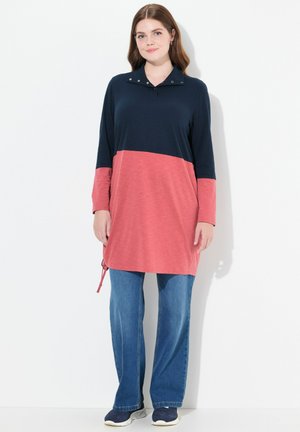 Langärmlige Tunika im Navy- und Korallenfarbblockdesign, mit Kragen, Druckknopfakzenten und Kordelzugdetail am Saum. Kombiniert mit ausgestellten Jeans.