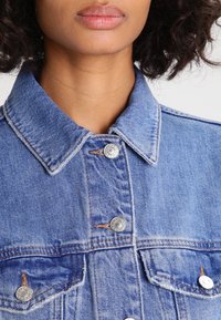 Veste en denim bleu clair avec un col pointu, des boutons-pression en argent, des poches poitrine et des détails de couture visibles. Tissu texturé.