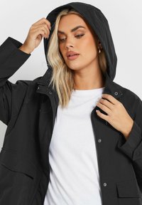 Svart huva jacka gjord av ett texturerat tyg, med tryckknappar, en oknäppt framsida och lapplommer. Bärs över en vit t-shirt.