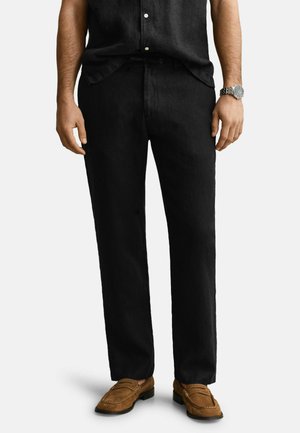 Homme portant un pantalon droit noir, des mocassins en daim marron, une chemise noire et une montre-bracelet en argent, debout sur un fond blanc.