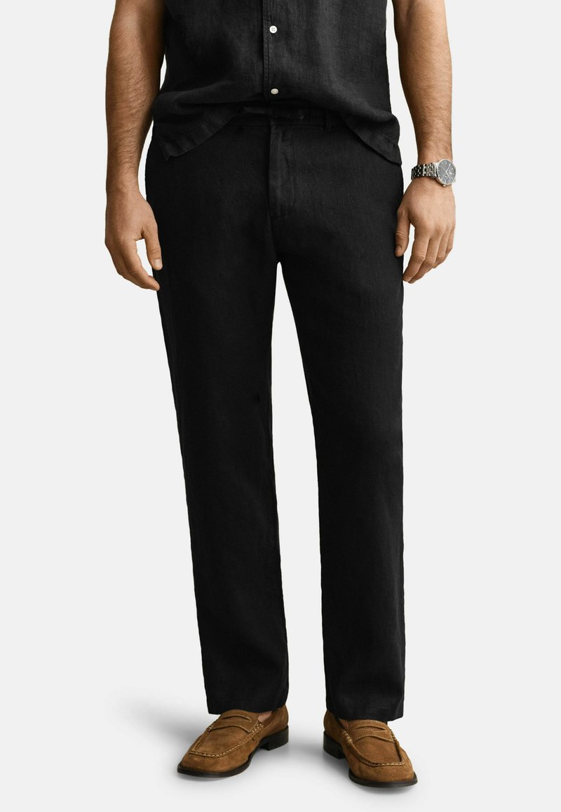 Mann trägt schwarze Hose mit geradem Bein, braune Wildleder-Loafer, schwarzes Hemd und silberne Armbanduhr, steht auf weißem Hintergrund.