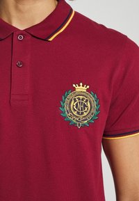 Burgundské polo tričko vyrobené z bavlny, s tříknoflíkovým zapínáním, vyšitým emblémem ve zlaté a zelené barvě a kontrastními pruhy na límečku.
