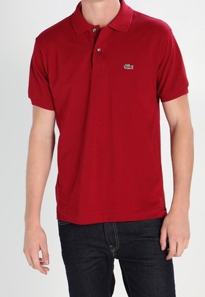 Mann trägt ein rotes Poloshirt mit kurzen Ärmeln und kleinem grünem Krokodil-Logo auf der Brust sowie dunkelblaue Jeans.