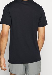 Hombre con una camiseta negra de manga corta lisa y pantalones cortos gris claro, visto desde atrás contra un fondo neutral.