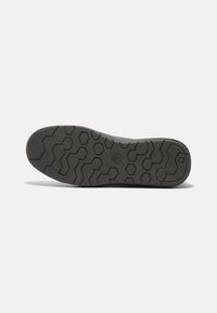 Suela de un zapato de color oscuro con patrones de diseño geométrico hexagonal y en zigzag, y un logo de árbol en el centro, sobre un fondo blanco.