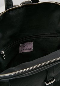 Interior de un bolso negro con un bolsillo con cremallera y una etiqueta de tela que muestra el nombre de la marca y el logo "Anna Field".