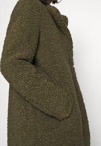 Primer plano de una persona que lleva abrigo de teddy texturizado color verde oliva, con la mano en el bolsillo lateral, arete de aro visible y barba, sobre un fondo blanco.