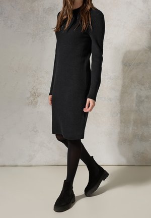 Kobieta w czarnej dzianinowej sukience z długim rękawem, czarnych rajstopach i czarnych botkach, stojąca na tle neutralnej, fakturowanej ściany.