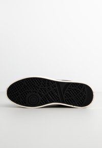 Schwarze Gummisneaker-Sohle mit strukturiertem Profil, umrandet von einem weißen Rand. Merkmale ein kreisförmiges Logo-Detail in der Nähe der Ferse.